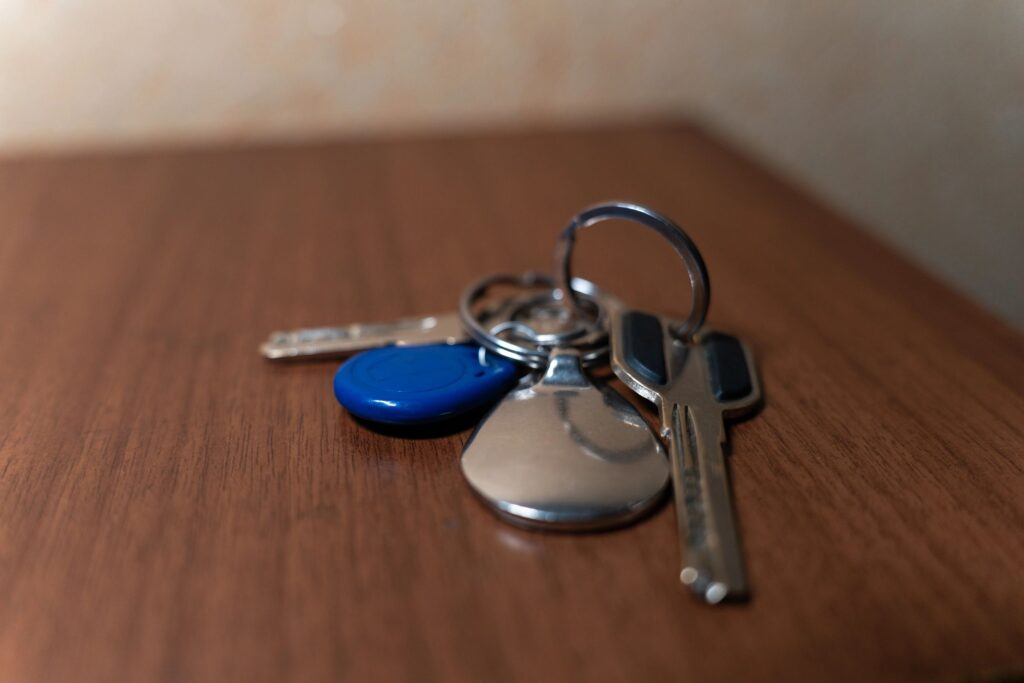 A set of metal keys with keychains and a blue fob lying on a wooden surface, with a red plastic tool blurred in the background. The image conveys everyday life, security, access, and household themes.
